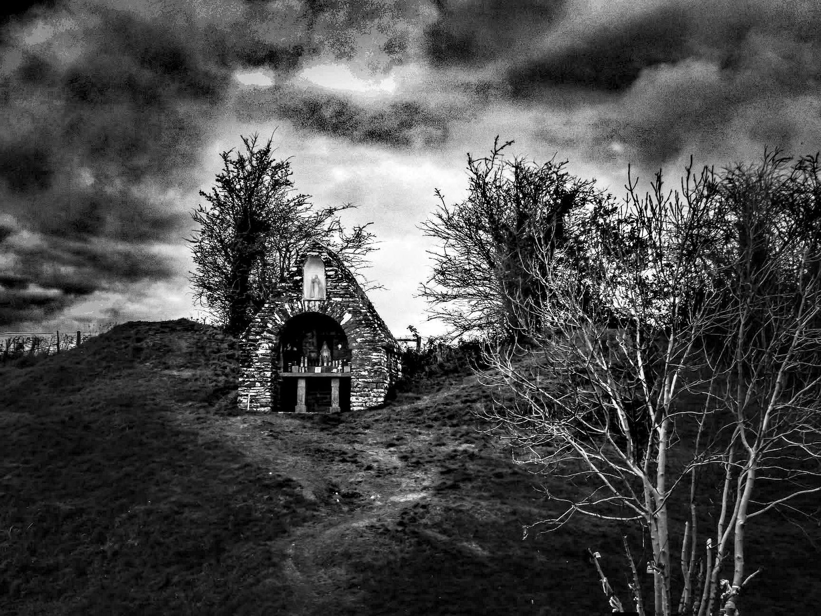 A black and white high-contrast photograph of St Ciarán's Holy Well and Shrine in Castlekeeran, County Meath, Ireland. The stone shrine sits on a grassy hillside under a dramatic, cloudy sky, with bare winter trees flanking the structure.